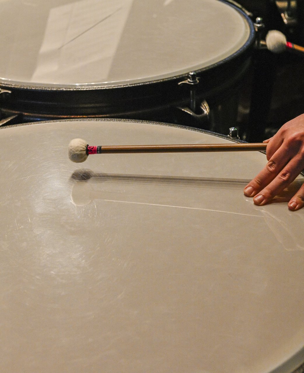 A hand resting a felt mallet on a timpani head, with more timpani behind